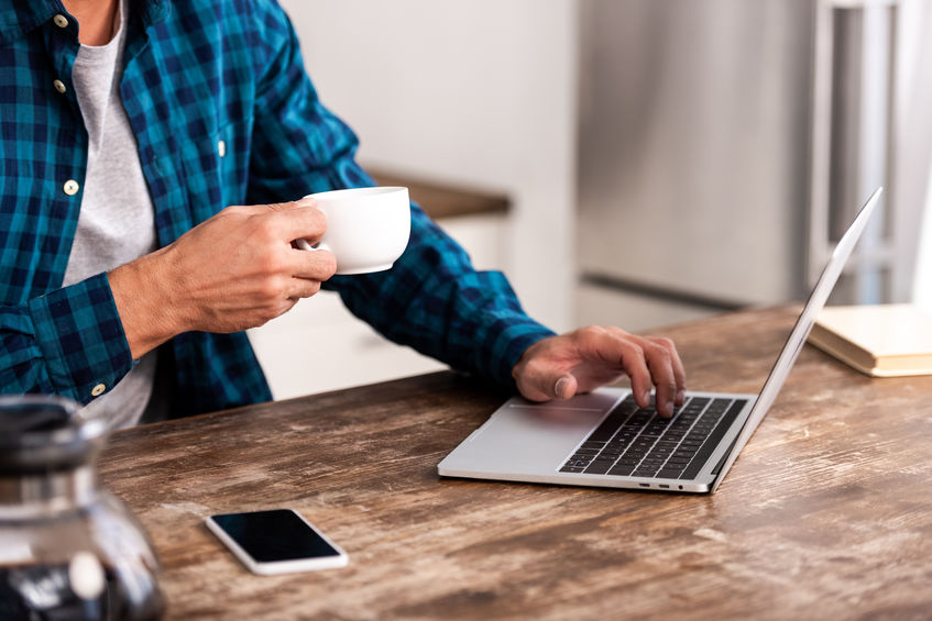 cropped shot of man holding cup of coffee and using laptop at home